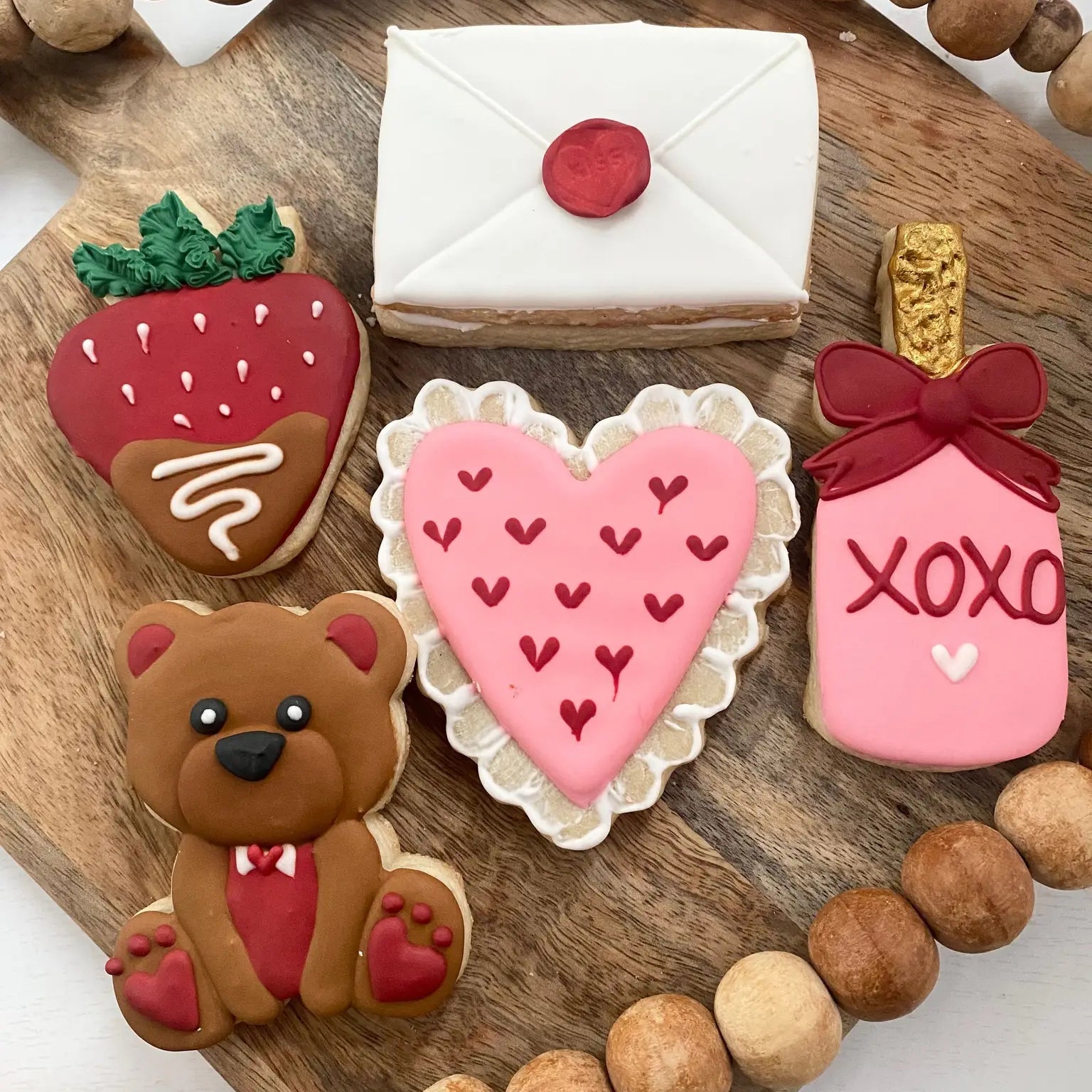 Decorative cookies shaped like a strawberry, envelope, heart, bear, and bottle with 'XOXO' on a wooden board.